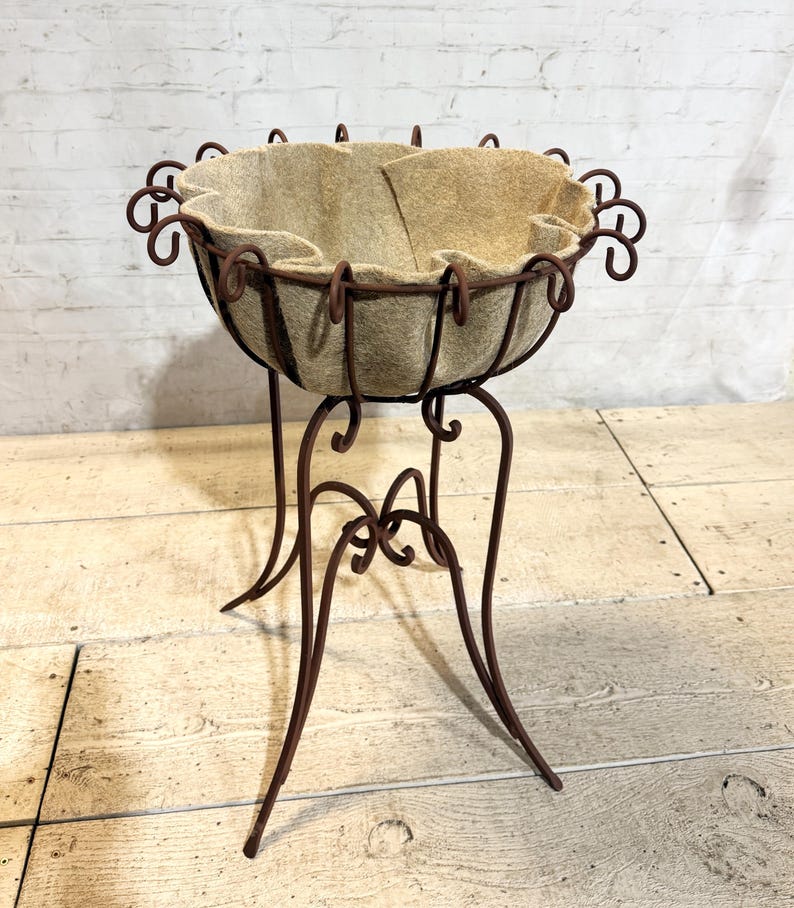 May include: A brown metal plant stand featuring a circular frame with decorative scrollwork accents and a tall, slender base. The bowl-shaped top is lined with a thick, beige felt fabric insert that conforms to the curved wire structure.