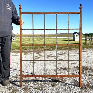 May include: A rusty metal trellis with a square grid pattern. The trellis is made of thick metal bars and has a rustic look. It is standing on a gravel surface.