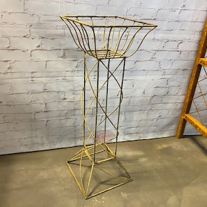 May include: A tall, ornate, metal plant stand with a square basket-like top. The stand has a geometric design with a twisted central column and a flared base. The metal has a distressed, aged gold finish, and the background is a white brick wall.