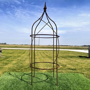May include: A tall, ornate, metal garden obelisk with a pointed top, set on a square of green grass. The obelisk has a circular mid-section and a base with three legs. The background features a grassy field, a blue sky, and small wooden structures.