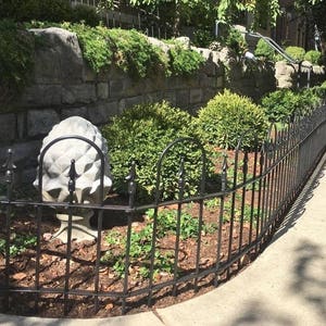 May include: A black metal fence with rounded tops surrounds a small garden area. A stone wall is behind the fence. A concrete pinecone sculpture is in the center of the garden.