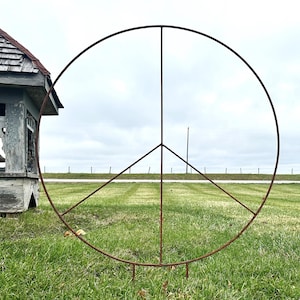 May include: A large, rusty metal peace sign stands in a grassy field. The sign is composed of a circle with three downward-pointing lines. In the background is a small, weathered gazebo and a cloudy sky.