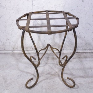 May include: A rustic, wrought iron plant stand with a circular top and a decorative, scrolled base. The stand has a grid pattern on the top and is made of dark, weathered metal.