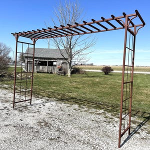May include: A rusty metal garden arbor with a trellis top and a lattice design on the sides. The arbor is standing on a gravel driveway.