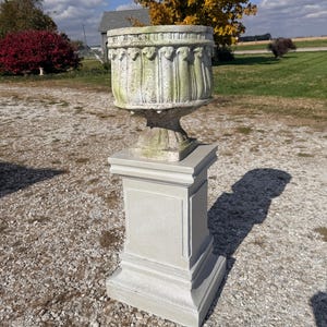 May include: A weathered, light gray stone planter with a decorative rim and vertical grooves, set atop a matching pedestal. The outdoor garden urn is in a rural setting with a gravel surface and trees in the background.