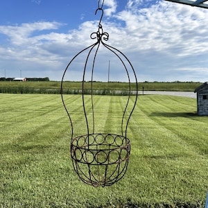 May include: A hanging metal planter with a hot air balloon design. The planter is made of dark brown metal and has a circular basket for holding plants. The background features a green lawn, a small wooden structure, and a blue sky.