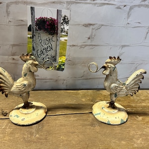 May include: Two white rooster-shaped metal picture holders with a ring to hold a photo or card. The roosters are standing on a wooden surface. The sign in the background reads "A Rustic Garden & Gifts".