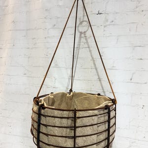 May include: A hanging planter featuring a cylindrical wire cage frame with a rusted brown finish. The interior is lined with a thick, light beige felt fabric. The structure is suspended by three thin metal rods that meet at a top ring.