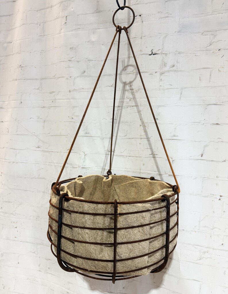 May include: A hanging planter featuring a cylindrical wire cage frame with a rusted brown finish. The interior is lined with a thick, light beige felt fabric. The structure is suspended by three thin metal rods that meet at a top ring.