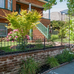 May include: A brick wall with a black metal fence in front of a house with a green porch and steps. There are green bushes and flowers in the yard.