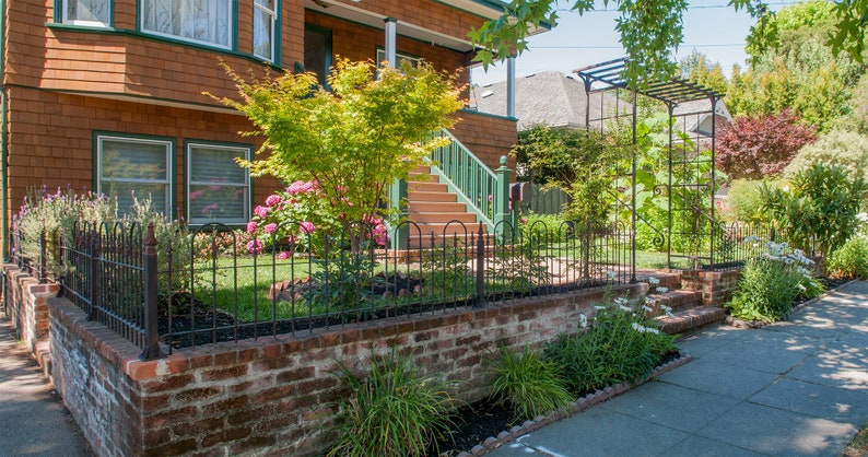 May include: A brick wall with a black metal fence in front of a house with a green porch and steps. There are green bushes and flowers in the yard.