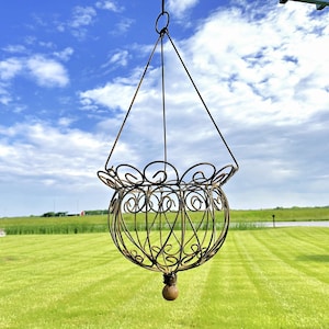 May include: A hanging, ornate metal basket with a spherical shape and decorative scrollwork. The basket is suspended by thin wires against a backdrop of a bright blue sky and green grass.
