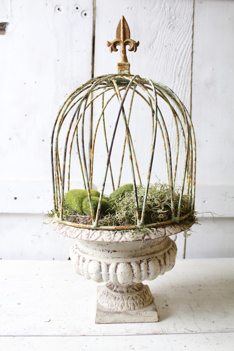 May include: A white ceramic planter with a decorative base and a wire cage top. The cage is decorated with a gold finial and filled with green moss.