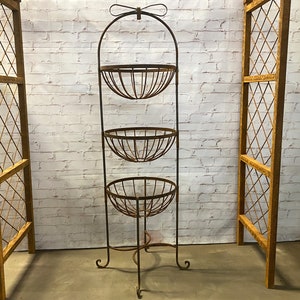 May include: A three-tiered, wrought-iron plant stand with three rounded baskets. The stand has a decorative bow at the top and sits on three curved legs. Two wooden trellis panels flank the stand, set against a white brick wall.