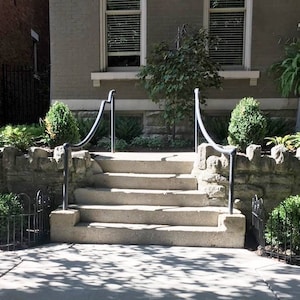 May include: A set of stone steps leading up to a house entrance. The steps are flanked by black metal railings. The steps are surrounded by a stone wall and lush greenery.