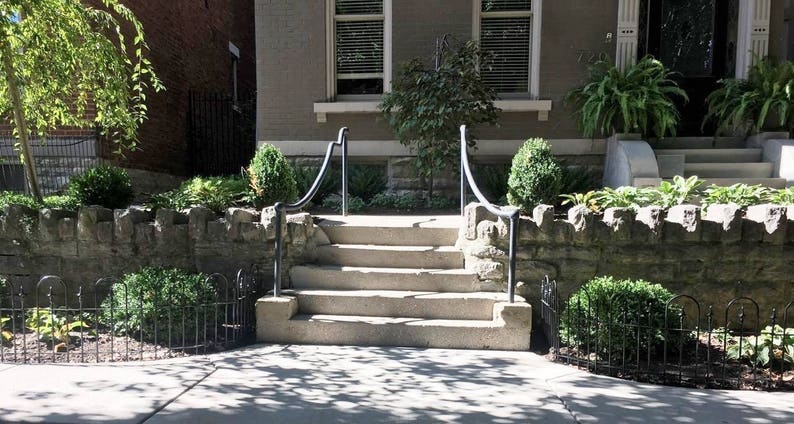 May include: A set of stone steps leading up to a house entrance. The steps are flanked by black metal railings. The steps are surrounded by a stone wall and lush greenery.