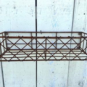 May include: A rectangular, rusty brown metal wire basket with a diamond pattern. The basket is mounted on a weathered, white-painted wooden surface. The basket is designed for wall mounting.