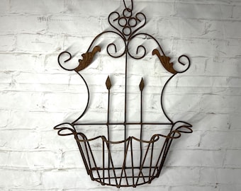 May include: A wall-mounted metal basket featuring a rusted brown finish, an open wire frame design, and decorative scrollwork at the top. The structure includes two vertical spear-shaped accents and a curved, basket-style base for storage.