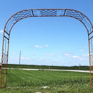 May include: A rustic metal archway with a decorative design, standing in a grassy field against a blue sky with white clouds.