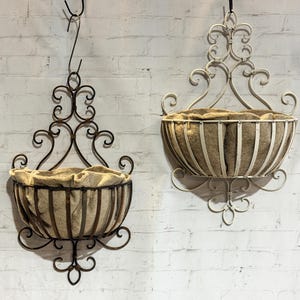 May include: Two wall-mounted metal baskets with scrollwork detailing and tan fabric liners. One basket is dark brown, and the other is off-white. Each basket features a semi-circular frame with vertical metal bars and an ornate, decorative backplate.
