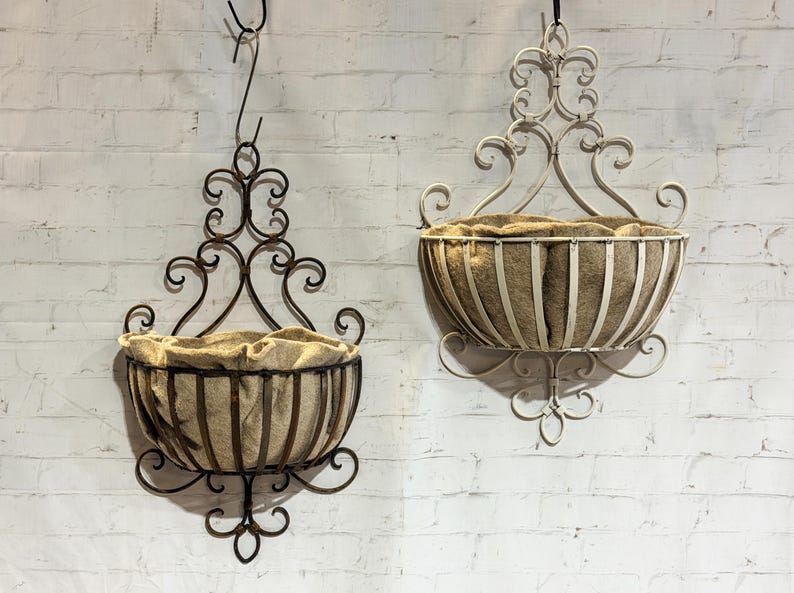 May include: Two wall-mounted metal baskets with scrollwork detailing and tan fabric liners. One basket is dark brown, and the other is off-white. Each basket features a semi-circular frame with vertical metal bars and an ornate, decorative backplate.