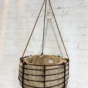 May include: A hanging planter featuring a dark brown, rusted metal wire frame basket with a beige felt liner inside. The basket is suspended by three thin metal rods connected to a top ring. A small white label inside reads 20 inches.