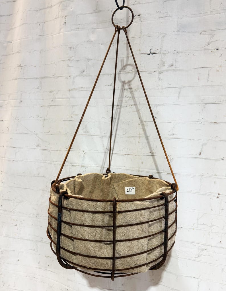 May include: A hanging planter featuring a dark brown, rusted metal wire frame basket with a beige felt liner inside. The basket is suspended by three thin metal rods connected to a top ring. A small white label inside reads 20 inches.