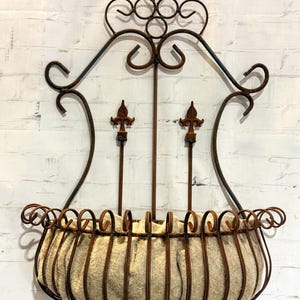 May include: A wall-mounted metal planter featuring a rust-colored finish with decorative scrollwork and two central fleur-de-lis accents. The semi-circular basket is constructed from vertical metal bars and contains a beige fiber liner.