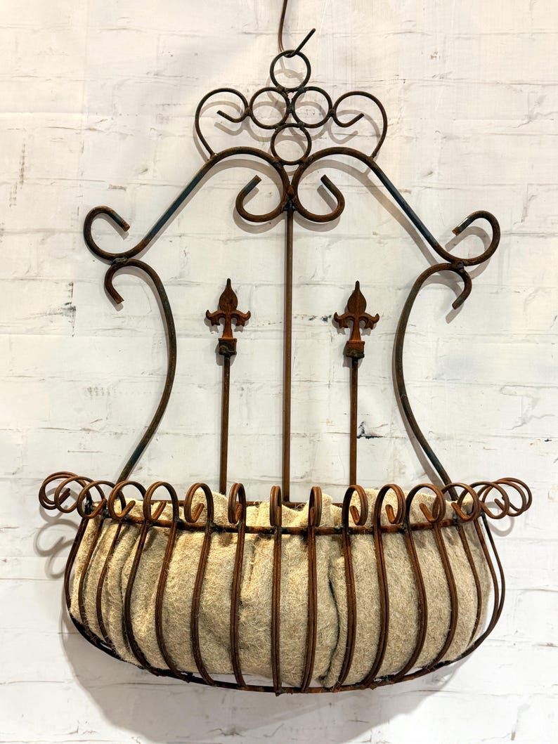 May include: A wall-mounted metal planter featuring a rust-colored finish with decorative scrollwork and two central fleur-de-lis accents. The semi-circular basket is constructed from vertical metal bars and contains a beige fiber liner.