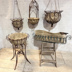 May include: A collection of five metal plant holders featuring dark brown wire frames with decorative scrollwork and beige fabric liners. Three are hanging baskets with chains, one is a pedestal planter, and one is a rectangular trough on a stand.