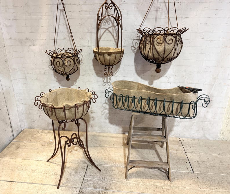 May include: A collection of five metal plant holders featuring dark brown wire frames with decorative scrollwork and beige fabric liners. Three are hanging baskets with chains, one is a pedestal planter, and one is a rectangular trough on a stand.