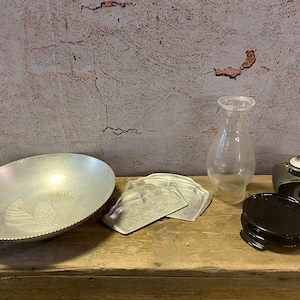 May include: A collection of vintage home decor items including a silver aluminum bowl with a raised design, a set of three silver aluminum coasters, a clear glass lamp chimney, a black ceramic base, and a black vintage camera.