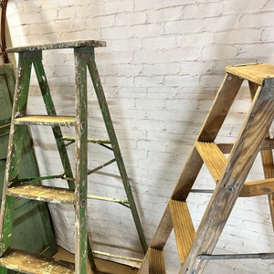 May include: Two wooden step ladders, one green and one natural wood, leaning against a white brick wall. Both ladders have three steps.