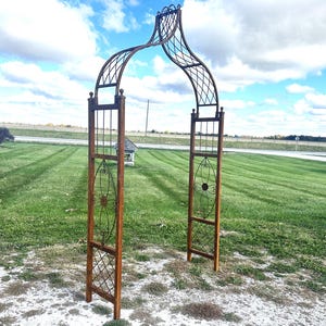 May include: A rustic metal garden archway with a decorative design. The archway is made of brown metal and has a curved top. It is standing on a grassy lawn.