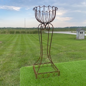 May include: A tall, ornate, wrought-iron plant stand with a weathered, rusty finish. The stand features a decorative, openwork basket-like top, supported by a slender, curved stem and a square base with circular accents. The stand is placed on a patch of green grass.