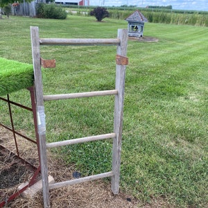 May include: A wooden ladder with four rungs and rusty metal brackets. The ladder is leaning against a metal fence with green artificial turf on top.