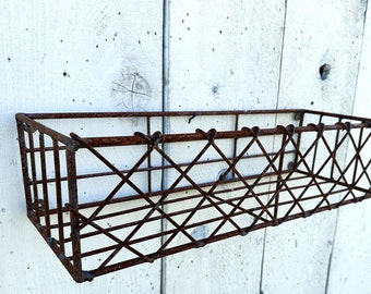 May include: A rectangular, rusty brown metal basket with a diamond pattern on the front and sides. The basket is mounted on a white wooden wall. This vintage-style basket is suitable for storage or display.