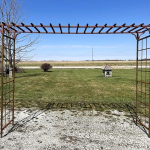 May include: A rusty metal garden arbor with a trellis top and a square lattice design on the sides. The arbor is standing on a gravel path in a grassy field.