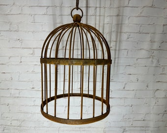Puede incluir: Una jaula de pájaros de metal oxidado con una parte superior abovedada y un lazo para colgar. La jaula está vacía y tiene un aspecto rústico y vintage.