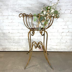 May include: An antique, wrought-iron plant stand with a basket-style top, filled with greenery and white flowers. The stand has a distressed, aged finish and decorative scrollwork. The legs are curved and splayed for stability, creating a vintage aesthetic.