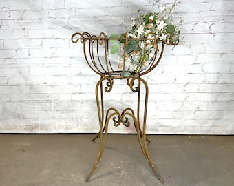 May include: An antique, wrought-iron plant stand with a basket-style top, filled with greenery and white flowers. The stand has a distressed, aged finish and decorative scrollwork. The legs are curved and splayed for stability, creating a vintage aesthetic.