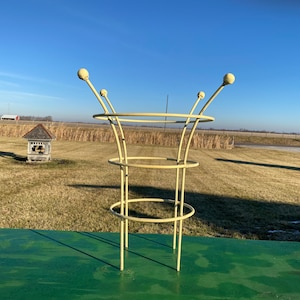May include: A pale yellow metal plant support cage featuring three circular rings connected by vertical rods, with each rod extending upward to terminate in a small spherical finial at the top of the structure.
