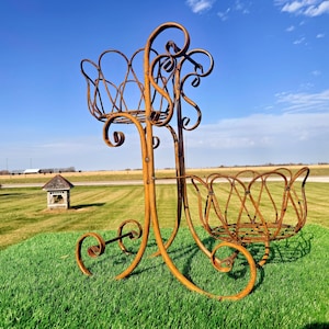 May include: A rusty brown metal plant stand with two basket-shaped holders. The stand has decorative scrollwork and sits on a green lawn under a blue sky. The plant stand is designed for outdoor use.