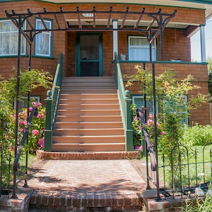May include: A brown wooden house with a white porch and a set of stairs leading up to the front door. The house has a green metal trellis over the porch and a lush green lawn.