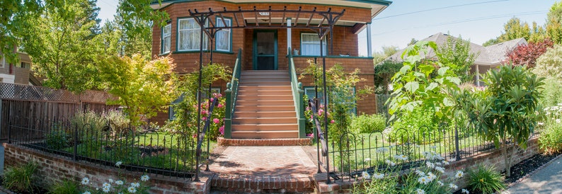 May include: A brown wooden house with a white porch and a set of stairs leading up to the front door. The house has a green metal trellis over the porch and a lush green lawn.