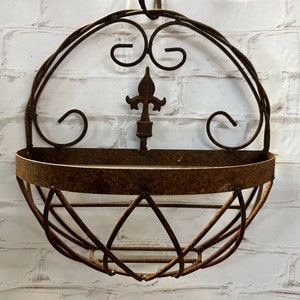 May include: A rustic, circular wall-mounted planter made of weathered, dark brown metal. The design features a curved top with decorative scrollwork, a central fleur-de-lis, and a basket-like lower section. The planter is designed to hold plants.