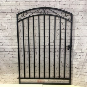 May include: Black metal gate with an arched top and decorative scrolls. The gate has vertical bars and a latch on the right side.