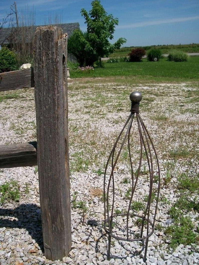 May include: A metal garden trellis with a round top and a decorative finial. The trellis is made of thin metal rods and is designed to support climbing plants.