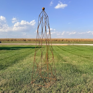 May include: A tall, decorative metal obelisk with a rust-colored finish stands in a grassy field under a blue sky. The obelisk has an open, geometric design and a spherical top. A small wooden structure is visible in the background.
