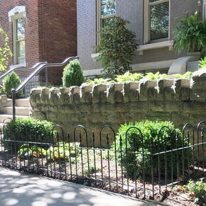 May include: A black metal fence with rounded top rails surrounds a small garden area in front of a brick building. The fence is in front of a stone wall and green bushes.